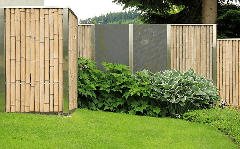 Full shot of large bamboo fence panels installed in a garden with large leafy plants and lawn in foreground